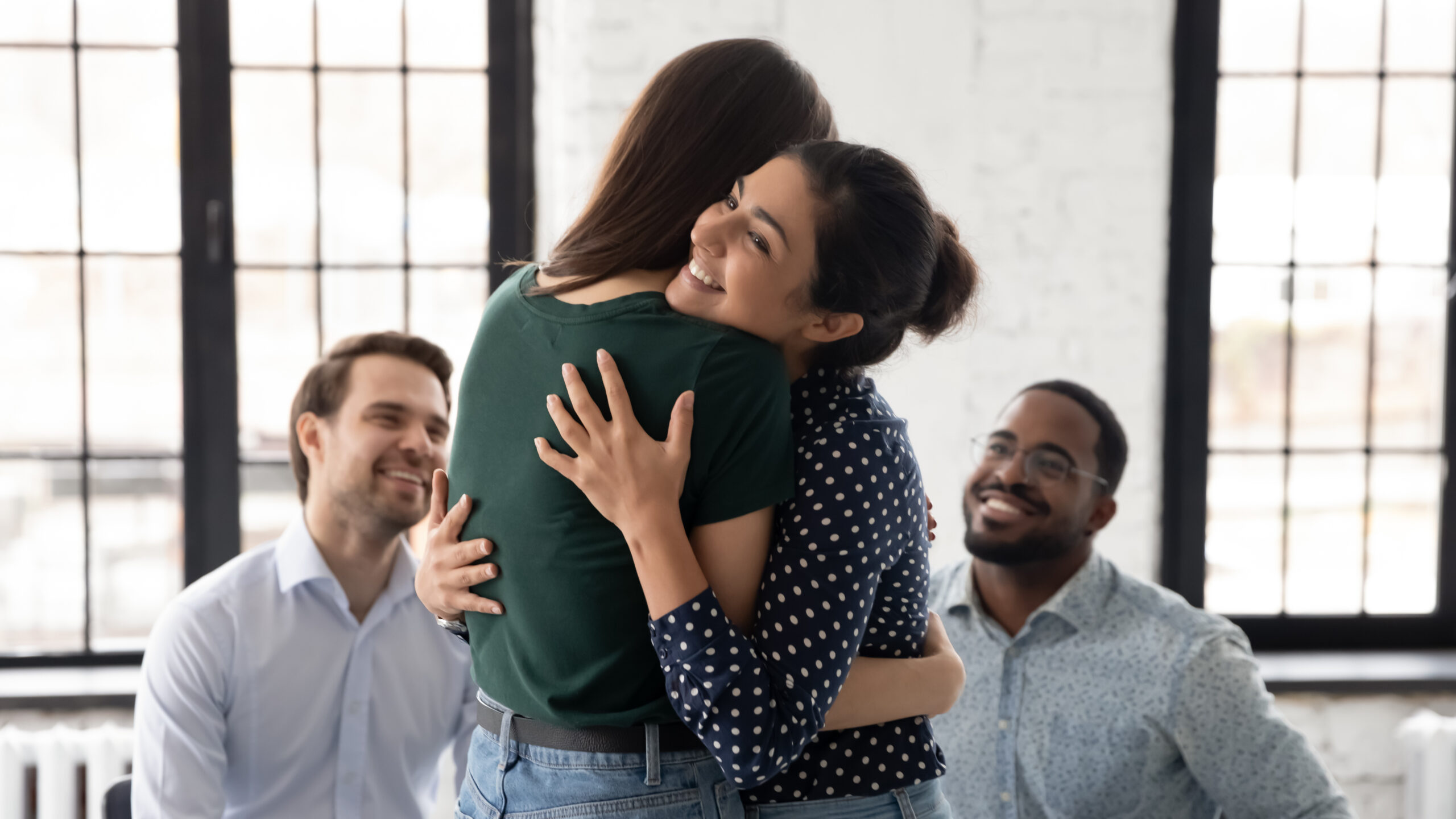 Two women embrace joyfully in a bright, airy room, while two smiling men look on, creating an atmosphere of celebration and camaraderie.