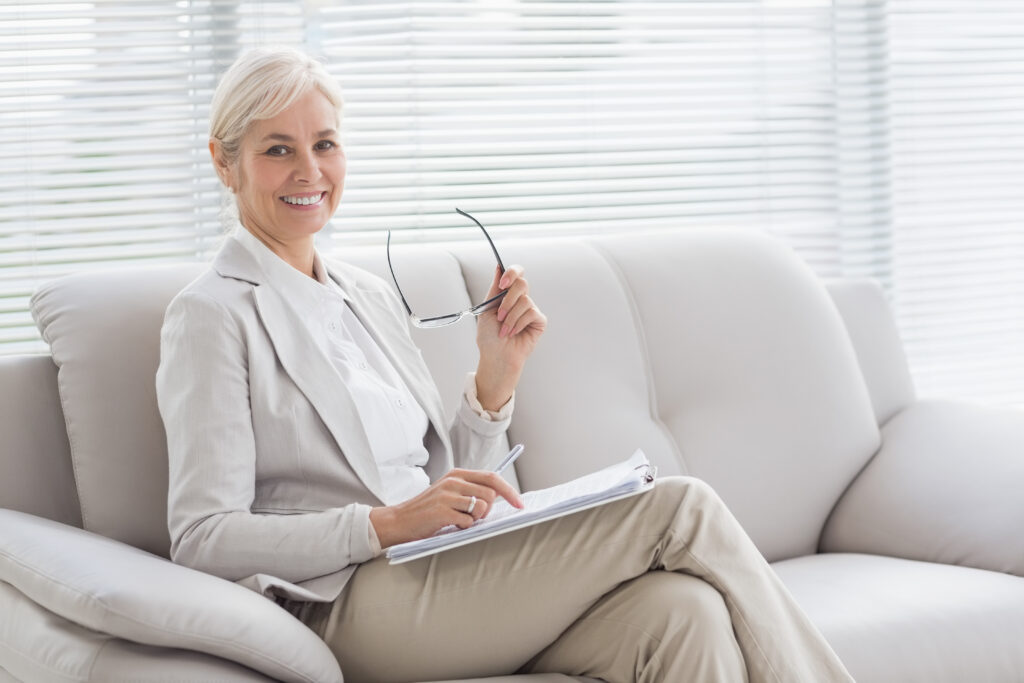 Smiling woman in a light suit sits on a sofa, holding glasses and a notebook, suggesting a professional or consultation setting.