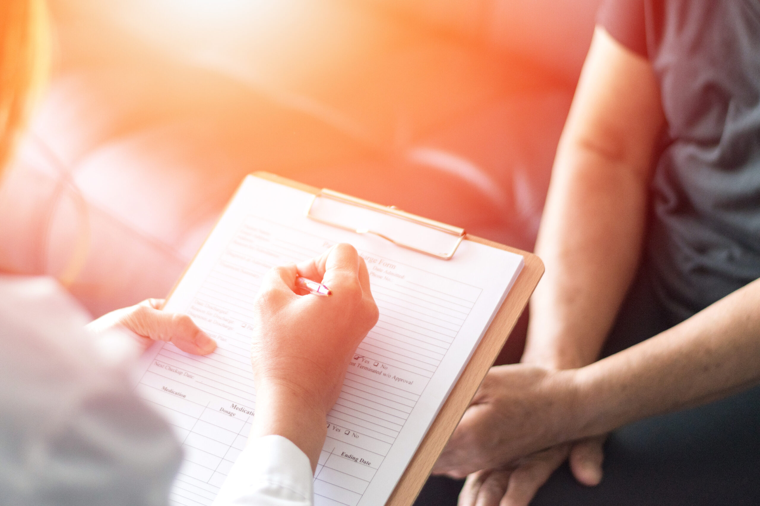 A healthcare professional writes on a clipboard while consulting with a patient