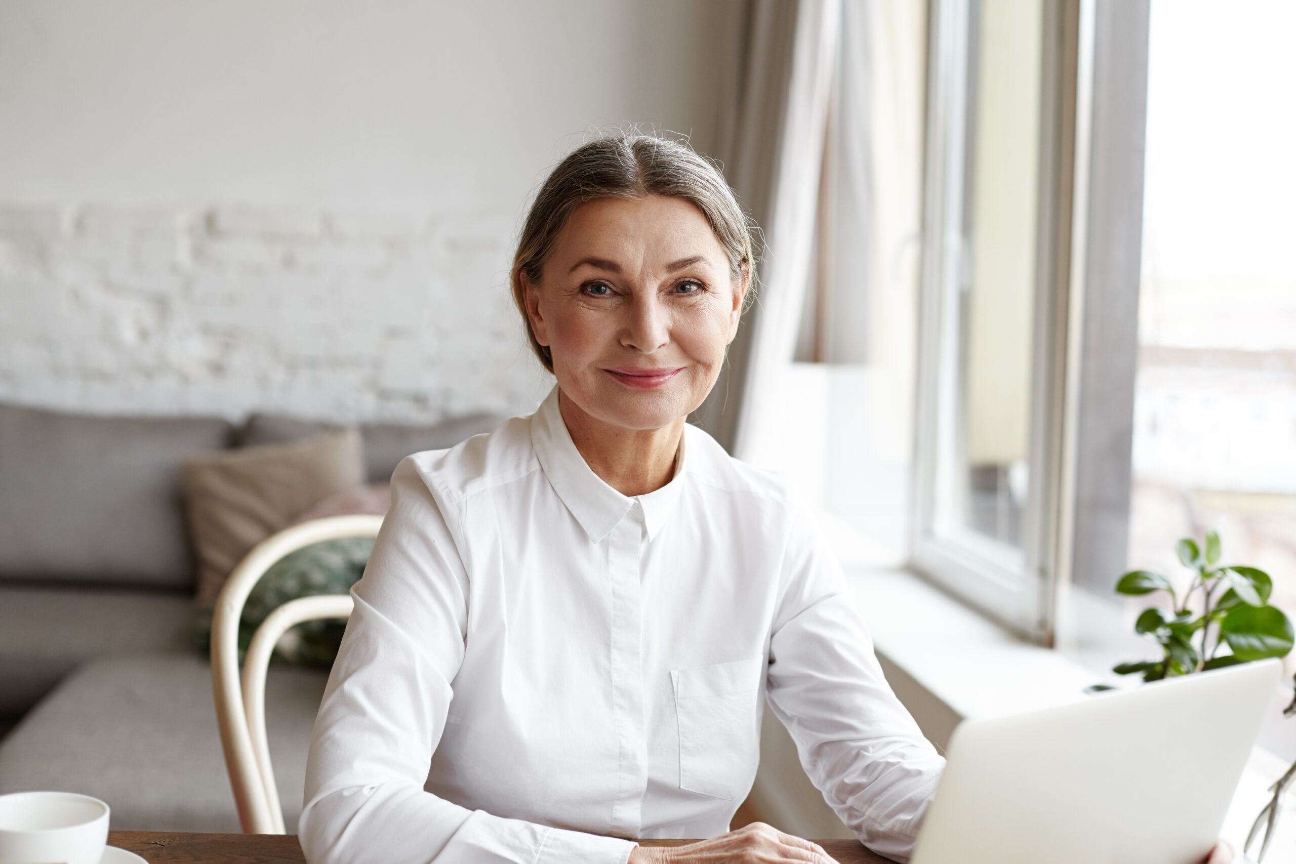Smiling woman in a white blouse sits at a wooden table with a laptop